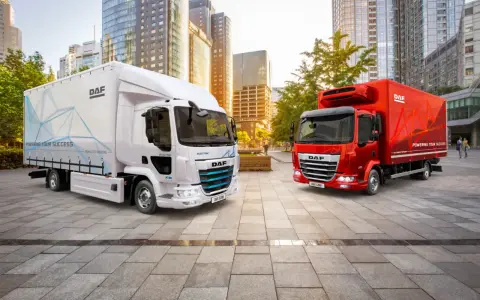 Dos camiones DAF, uno blanco eléctrico y otro rojo, en una ciudad moderna. El DAF XB blanco muestra "POWERING YOUR SUCCESS".