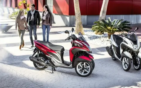 Dos motocicletas Yamaha Tricity, una roja y otra blanca, listas para circular. Tres personas pasean al fondo.