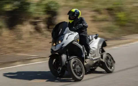 Un motorista con casco amarillo y negro conduce una moto de tres ruedas por una carretera. La moto ciudad se mueve rápido, creando un efecto de desenfoque.