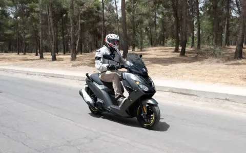 Hombre en casco y chaqueta conduce un triciclo de motor gris por una carretera rodeada de pinos.
