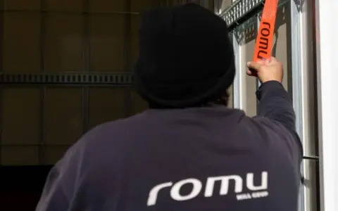 Persona con gorro negro ajusta una cinta naranja con el logo "romu", evitando una multa por exceso de peso.