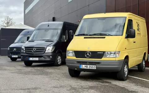 Tres furgonetas Mercedes-Benz Sprinter, una amarilla conmemorativa de 30 años, una negra y una azul, aparcadas frente a un edificio moderno.
