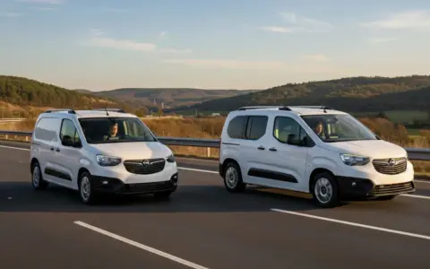 Dos furgonetas panel blancas circulan por una carretera con colinas boscosas al fondo.