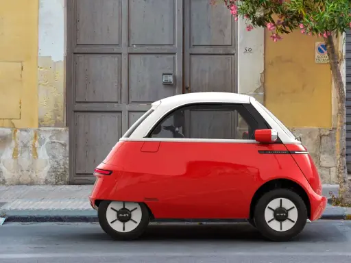 Un triciclo y cuadriciclo de motor rojo y blanco, un vehículo eléctrico compacto, estacionado frente a una puerta de madera y una pared amarilla.