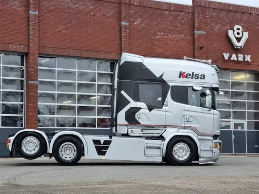 Camión Scania Longline plateado con diseño negro y rojo, aparcado frente a un edificio de ladrillo con puertas de garaje.