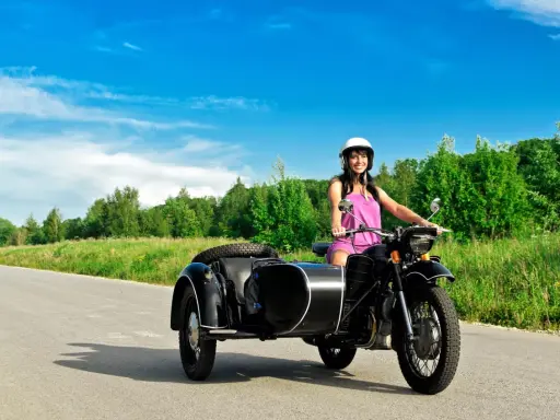 Mujer sonriente con casco blanco conduce una motocicleta negra con sidecar. El permiso A1 autoriza a conducir motocicletas con sidecar.