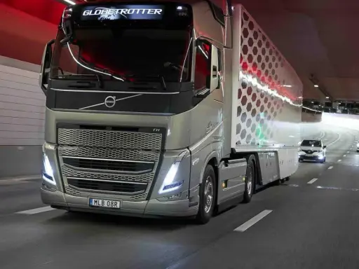 Camión Volvo FH Globetrotter plateado avanza por un túnel con luces rojas. El camión, que parece ser un modelo i-Save, transporta una carga con un diseño hexagonal.