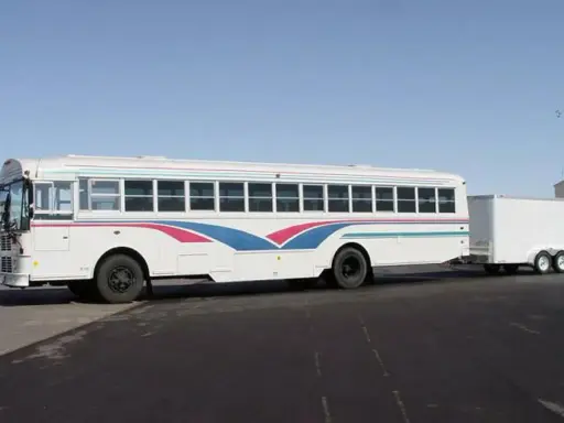 Autobús blanco con franjas azules y rosas, remolcando un tráiler. Ideal para un viaje largo con el carnet D+E.