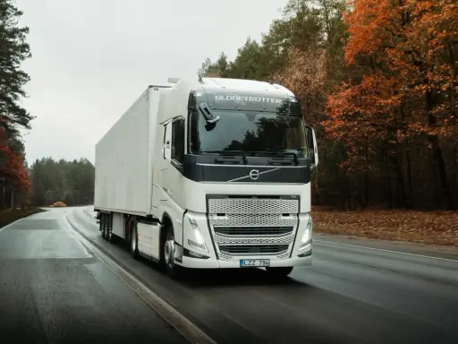 Un camión Volvo FH blanco con remolque blanco circula por una carretera mojada rodeada de árboles otoñales.