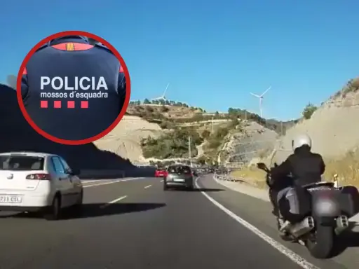 Un motorista de los Mossos d'Esquadra circula por el arcén de una autopista, adelantando a otros vehículos en un día soleado.