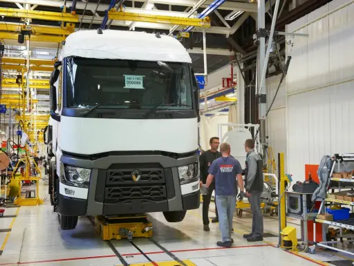 Tres trabajadores observan un camión Renault en una línea de montaje. El camión está sobre una plataforma amarilla.