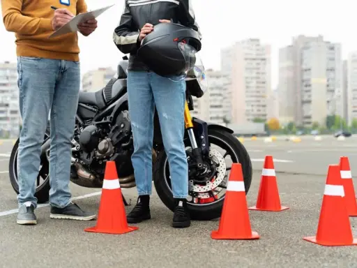 Persona se prepara para el circuito examen moto a1. Un instructor revisa una lista junto a una moto y conos naranjas.
