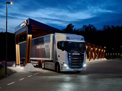 Camión Scania eléctrico en una estación de carga nocturna. El remolque dice "TAKE CHARGE 100% ELECTRIC".