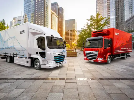Dos camiones DAF, uno blanco eléctrico y otro rojo, en una ciudad moderna. El DAF XB blanco muestra "POWERING YOUR SUCCESS".