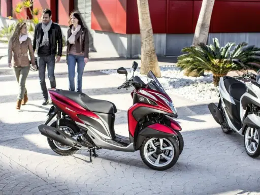 Dos motocicletas Yamaha Tricity, una roja y otra blanca, listas para circular. Tres personas pasean al fondo.