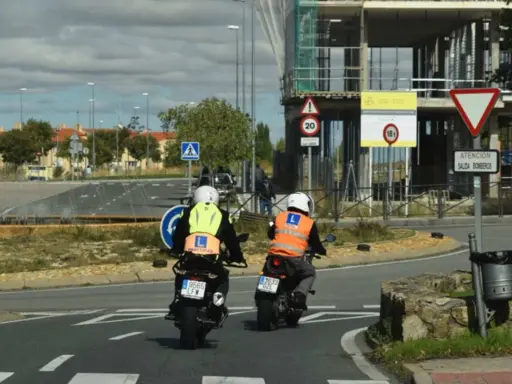 Dos motoristas con chalecos de prácticas, uno amarillo y otro naranja, circulan en moto. Uno de ellos lleva un cartel de "carnet moto 300".