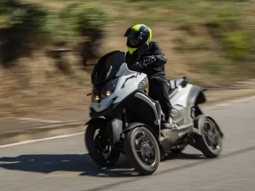 Un motorista con casco amarillo y negro conduce una moto de tres ruedas por una carretera. La moto ciudad se mueve rápido, creando un efecto de desenfoque.