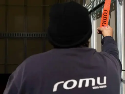 Persona con gorro negro ajusta una cinta naranja con el logo "romu", evitando una multa por exceso de peso.