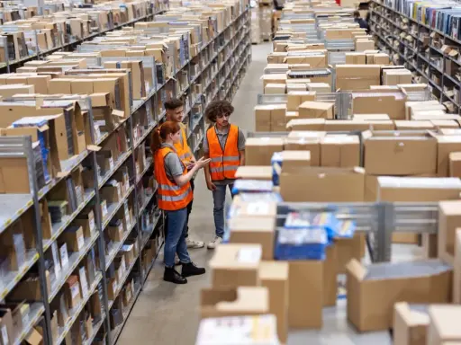 Tres trabajadores revisan inventario en un almacén, optimizando la logística de aprovisionamiento. Estanterías llenas de cajas y productos se extienden hasta el fondo.