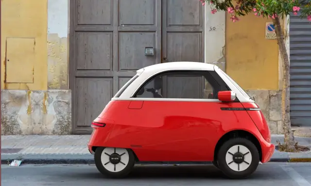 Un triciclo y cuadriciclo de motor rojo y blanco, un vehículo eléctrico compacto, estacionado frente a una puerta de madera y una pared amarilla.