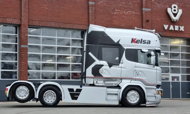 Camión Scania Longline plateado con diseño negro y rojo, aparcado frente a un edificio de ladrillo con puertas de garaje.
