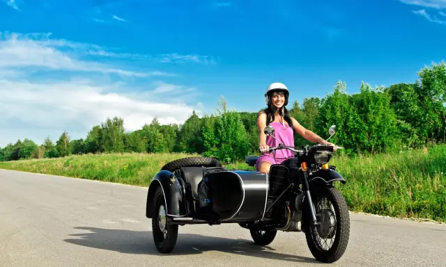 Mujer sonriente con casco blanco conduce una motocicleta negra con sidecar. El permiso A1 autoriza a conducir motocicletas con sidecar.