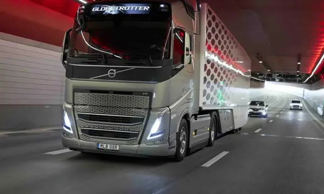 Camión Volvo FH Globetrotter plateado avanza por un túnel con luces rojas. El camión, que parece ser un modelo i-Save, transporta una carga con un diseño hexagonal.