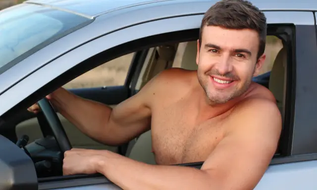 Hombre sonriente al volante de un coche, disfrutando de la libertad de que se puede conducir sin camiseta en un día caluroso.