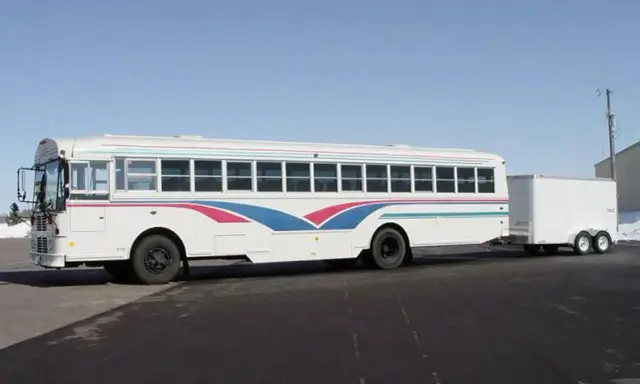 Autobús blanco con franjas azules y rosas, remolcando un tráiler. Ideal para un viaje largo con el carnet D+E.