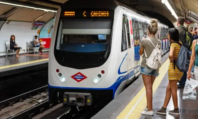 En la estación de metro, un tren blanco espera. Varias personas observan, una podría recibir una multa por usar tarjeta de transporte de otra persona.