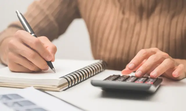 Calculando el precio de clases de conducir. Mano con bolígrafo en cuaderno y otra mano en calculadora.