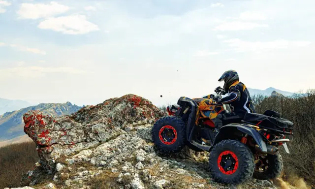 Un piloto con casco y chaqueta amarilla y negra conduce un quad ATV por un terreno rocoso y montañoso. El permiso para esta aventura es la emoción.