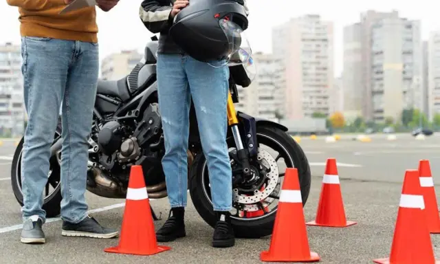 Persona se prepara para el circuito examen moto a1. Un instructor revisa una lista junto a una moto y conos naranjas.