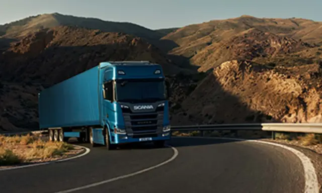 Camión Scania azul recorre una carretera sinuosa entre montañas áridas, evocando la fuerza y el espíritu de aventura de Suecia.