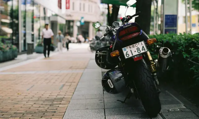 Una moto Yamaha aparcada en la acera, con dos cascos colgados. Al fondo, gente pasea por la calle.