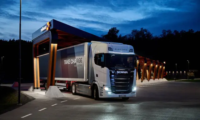 Camión Scania eléctrico en una estación de carga nocturna. El remolque dice "TAKE CHARGE 100% ELECTRIC".