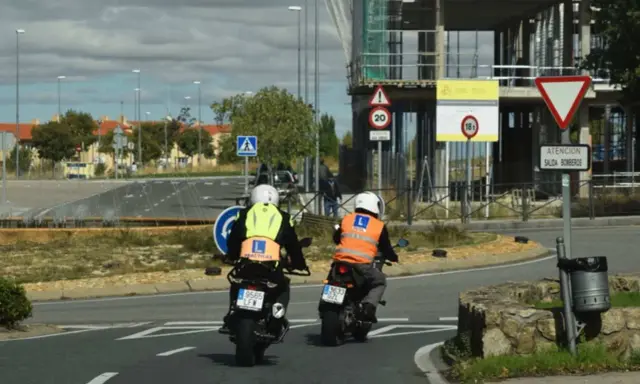 Dos motoristas con chalecos de prácticas, uno amarillo y otro naranja, circulan en moto. Uno de ellos lleva un cartel de "carnet moto 300".