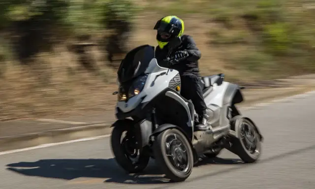 Un motorista con casco amarillo y negro conduce una moto de tres ruedas por una carretera. La moto ciudad se mueve rápido, creando un efecto de desenfoque.