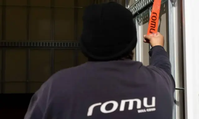 Persona con gorro negro ajusta una cinta naranja con el logo "romu", evitando una multa por exceso de peso.