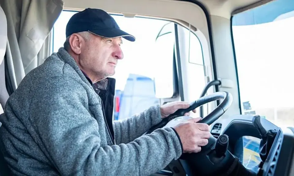 Conductor de camión con gorra y chaqueta gris, con las manos en el volante. Podría ser parte de un listado de cese de actividad de transportistas.