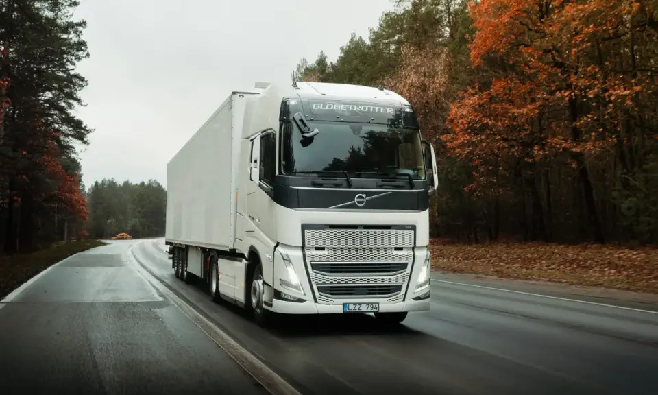 Un camión Volvo FH blanco con remolque blanco circula por una carretera mojada rodeada de árboles otoñales.