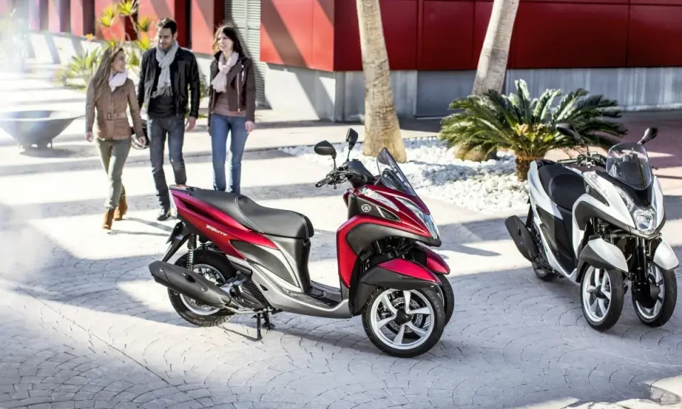 Dos motocicletas Yamaha Tricity, una roja y otra blanca, listas para circular. Tres personas pasean al fondo.