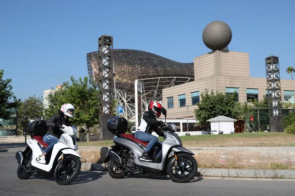 Dos personas en moto ciudad, con cascos, circulan por una calle con edificios modernos y un cielo azul.