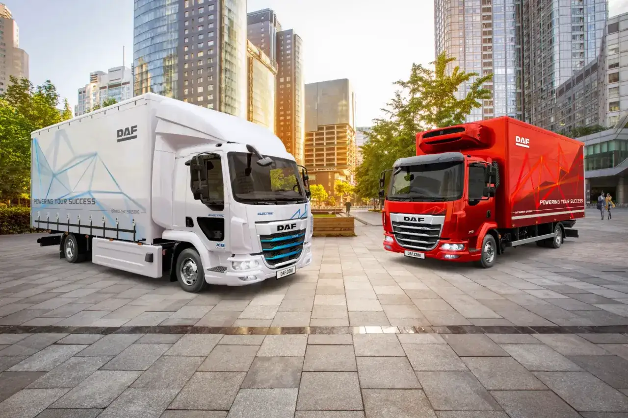Dos camiones DAF, uno blanco eléctrico y otro rojo, se exhiben en un entorno urbano moderno. El DAF XB blanco destaca por su diseño futurista y el lema 