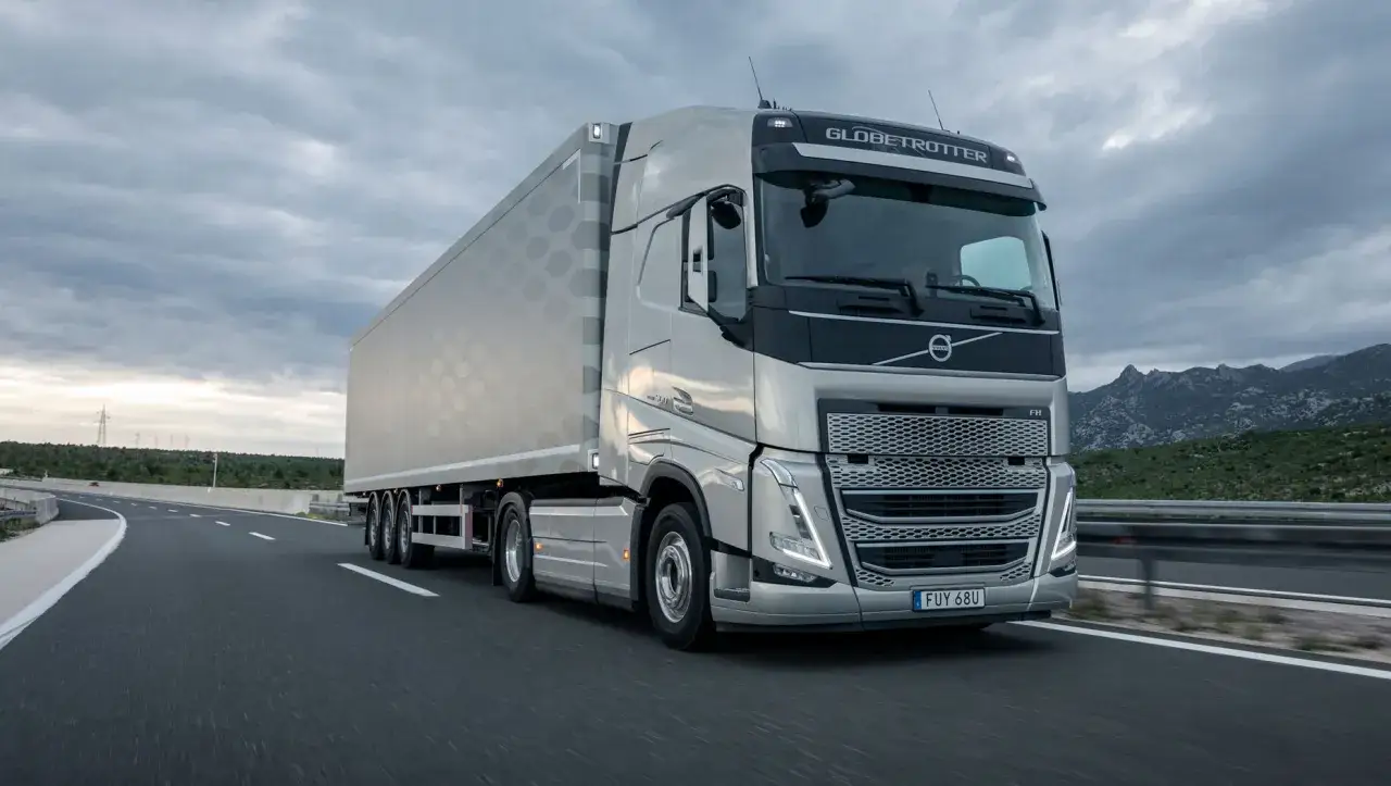 Un camión Volvo FH plateado con remolque avanza por una carretera con montañas al fondo y un cielo nublado.