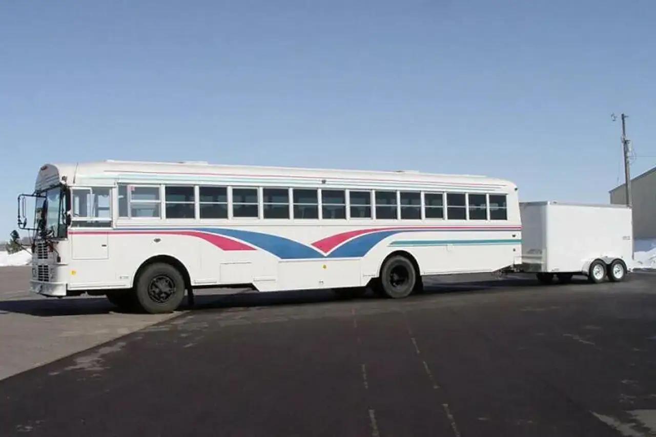 Autobús blanco con franjas azules y rosas, remolcando un tráiler. Ideal para quienes poseen el carnet D+E y buscan versatilidad.
