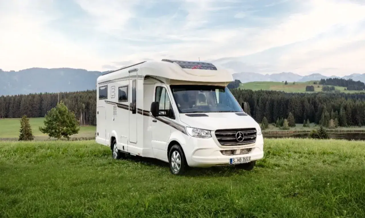 Autocaravana blanca de más de 3500 kg aparcada en un prado verde con montañas y bosque al fondo.
