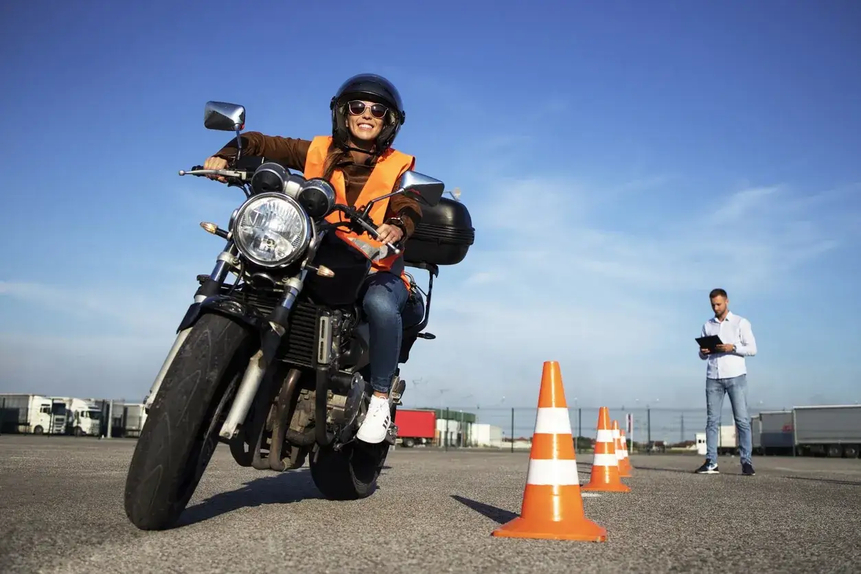 Mujer sonriente con chaleco naranja y casco en moto, lista para obtener el carnet A2. Instructor observa.