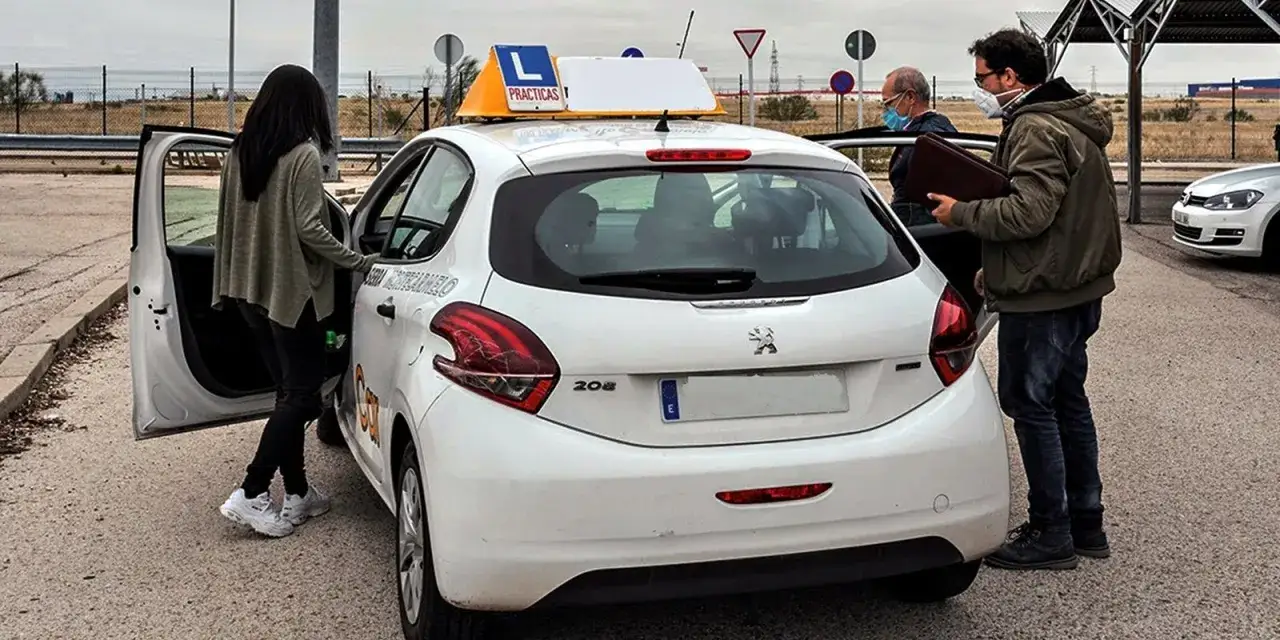 Persona se prepara para examen de conducir. El instructor, con mascarilla, revisa documentos. Se ven se&ntilde;ales de tr&aacute;fico y el coche de pr&aacute;cticas con la 