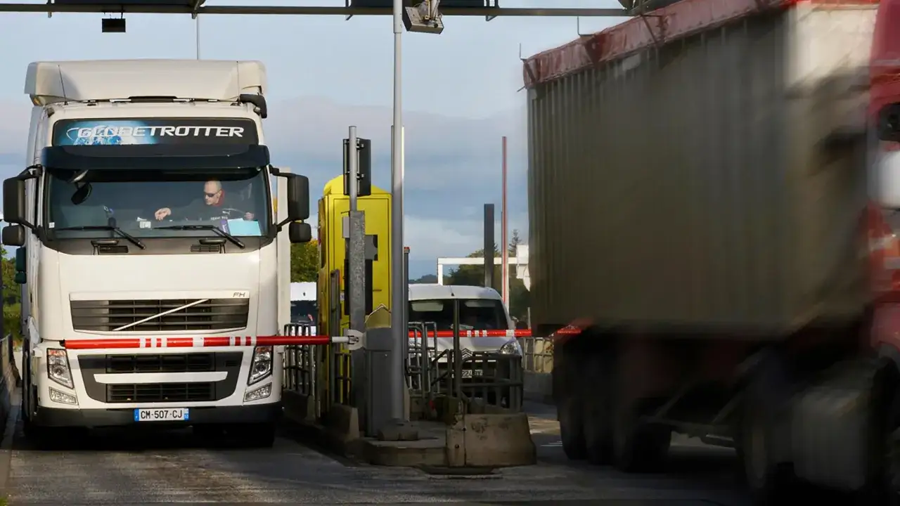Camión blanco en peaje, listo para cruzar a Luxemburgo o Holanda. Otro camión pasa borroso.